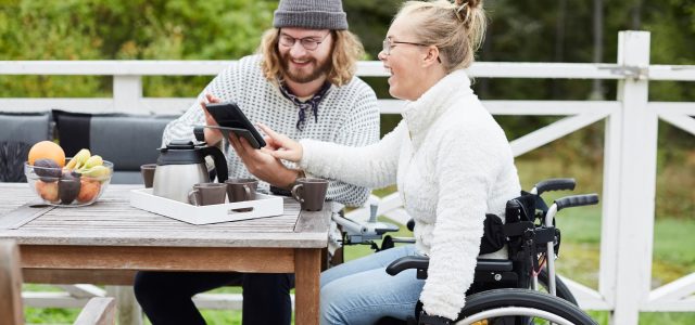 Manlig vårdgivare och funktionshindrad kvinna sitter vid bord på altan och använder surfplatta tillsammans