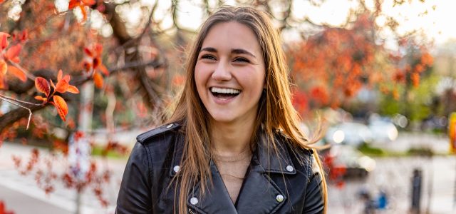 Cool and funny young smiling woman in urban setting surrounded by red leafy trees at sunset
